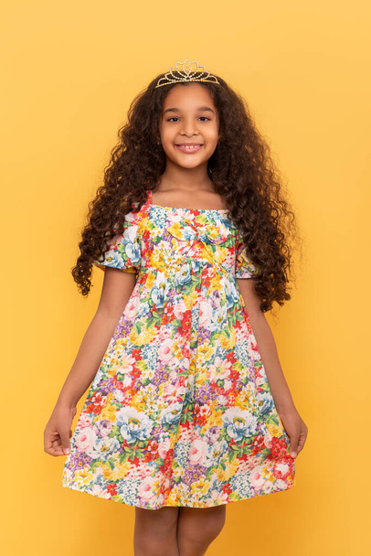 Half length portrait of a dark skinned young girl with long curly hair with a crown on his head against yellow background - Foto, Imagen