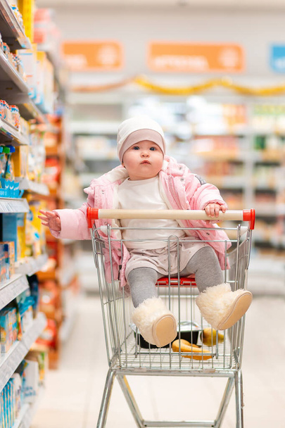 Ostoksilla. Söpö lapsi istuu ostoskärryissä keskellä supermarketia. Pystysuora. Perheostosten ja vanhemmuuden käsite - Valokuva, kuva