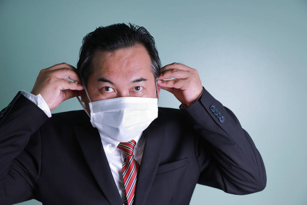 Der asiatische Geschäftsmann trägt einen schwarzen Anzug und blickt in die Kamera. Zwei Hände tragen medizinische Masken, um dem Coronavirus vorzubeugen. Gesundheitskonzept schützt Covid-19. - Foto, Bild