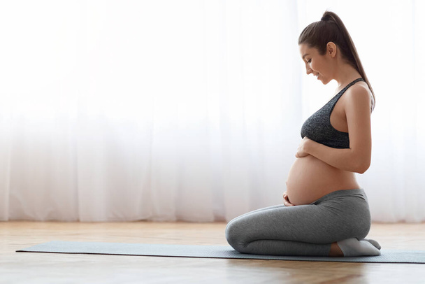 Terhes nő edzés jóga otthon, ül a szőnyegen, és megérinti has - Fotó, kép