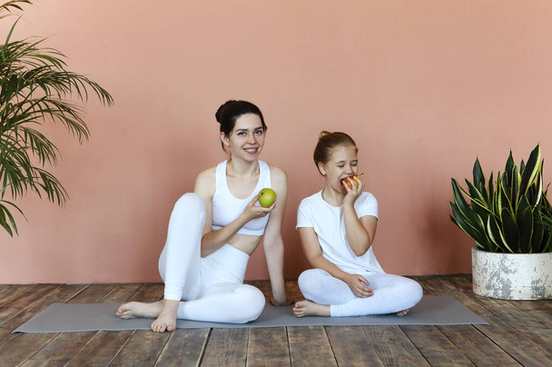 Glimlachende vrouw en klein meisje eten fruit na het beoefenen van yoga op mat in gezellige woonkamer  - Foto, afbeelding