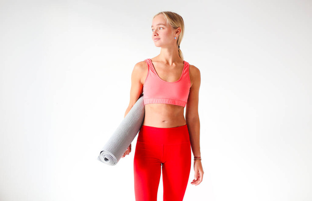 Young slim female in bright red top and leggings holding mat for yoga practice in white studio - Photo, Image