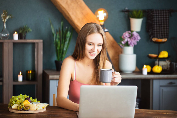 Joyeux jeune femme avec tasse de boisson chaude souriant et ordinateur portable de navigation le matin à la maison - Photo, image