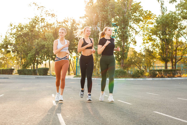 Parktaki güneşli bir günde asfalt otoparkta gülümseyen ve koşan genç sporcu kadınlar. - Fotoğraf, Görsel