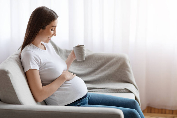 Улыбающаяся молодая беременная женщина отдыхает на диване с чашкой травяного чая - Фото, изображение