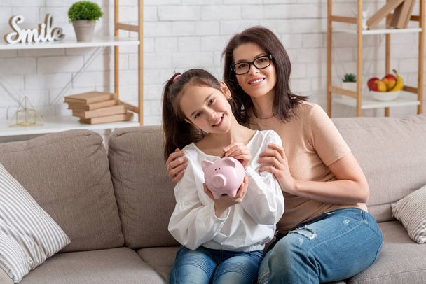 Happy young mother and her teen daughter holding piggybank, sitting on sofa at home, copy space - Photo, Image