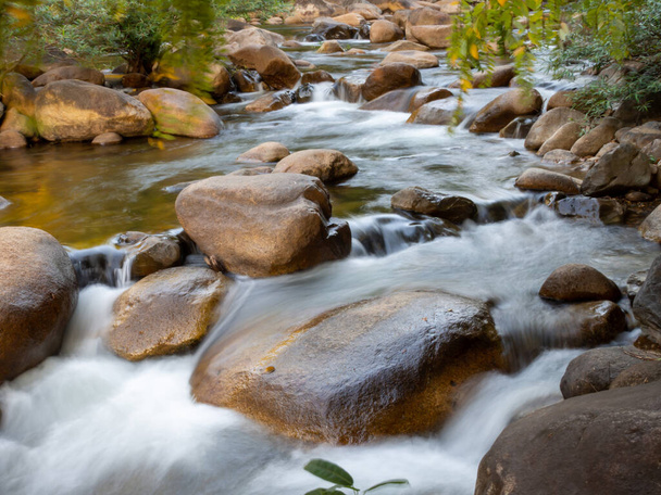 Eine Langzeitaufnahme eines kleinen Baches, der durch einen braunen Felsen fließt. Und es gibt grüne Bäume als Hintergrund. Es ist ein spezieller Schwerpunkt. - Foto, Bild