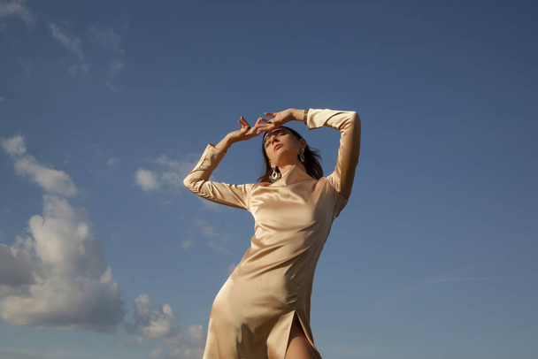 Fashion outdoor portret van vrouw in beige zijde satijn lange jurk tegen heldere blauwe lucht - Foto, afbeelding