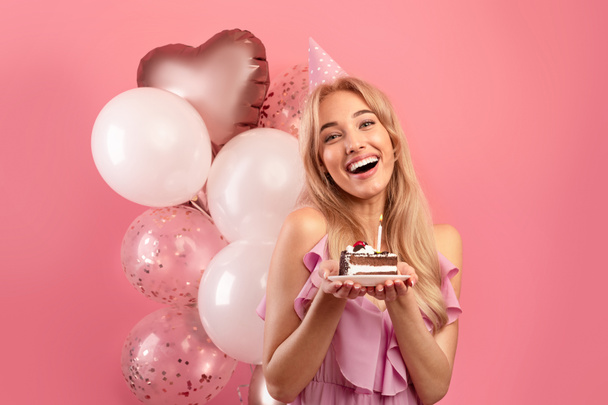 Mooie blonde vrouw met verjaardagshoed, ballonnen en lekkere taart kijkend naar camera en lachend op roze achtergrond - Foto, afbeelding