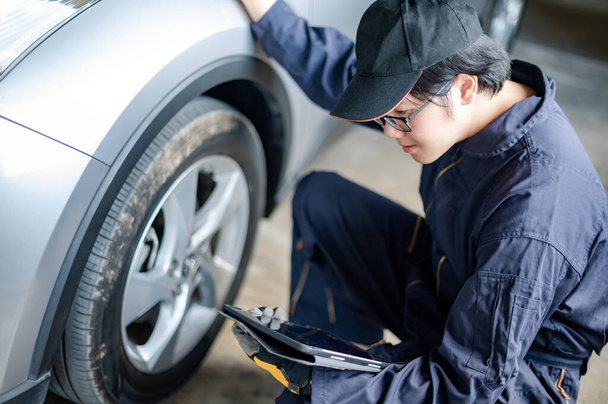 アジアの自動車整備士は、自動車サービスガレージでデジタルタブレットチェック車ホイールとタイヤを保持しています。自動車産業で働く機械メンテナンスエンジニア。自動車サービスと修理の概念 - 写真・画像