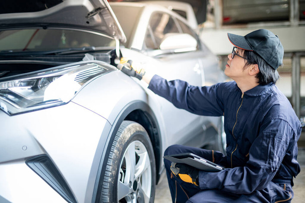 Asian auto mechanic holding digital tablet checking car wheel and tire in auto service garage. Mechanical maintenance engineer working in automotive industry. Automobile servicing and repair concept - 写真・画像