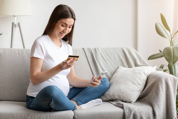 Happy pregnant woman shopping online, using smartphone and credit card at home, - 写真・画像