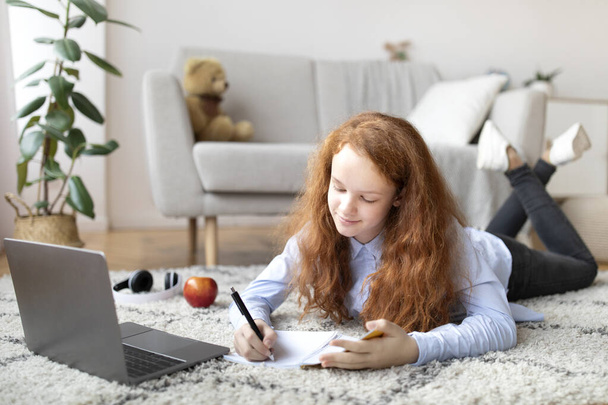Mädchen liegt auf Teppich, benutzt Laptop, schreibt in Notizbuch - Foto, Bild