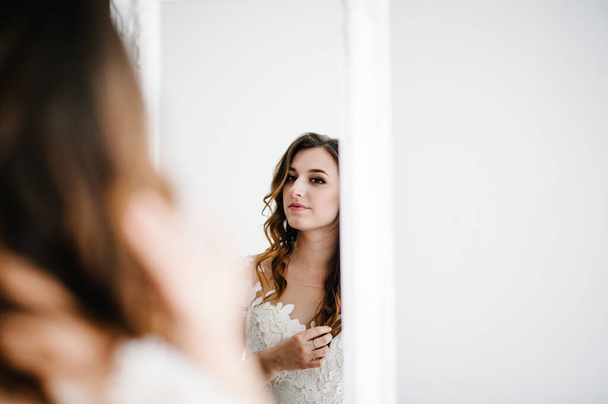 Young attractive smiling bride in dress stand in front of the mirror and looking. Reflection.  Portrait woman. The concept of the holiday and love. Wedding Morning Preparations. - Fotó, kép