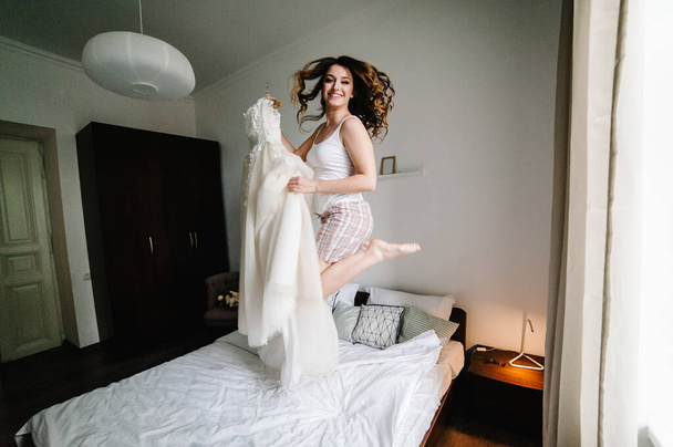 The bride holds a wedding dress in her hands and spins, turn and jumping on the bed. Style vintage elegant dress with lace. Wedding Morning Preparations. - Foto, Imagem