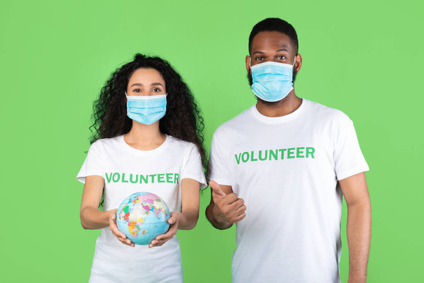 Волонтеры, несущие маски, держащие в руках глобус, поднимая пальцы вверх, зеленый фон - Фото, изображение