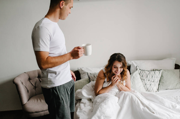 Milující manžel nosí kávu pro svou šťastnou ženu v posteli, ve velké ložnici, boční pohled. Valentýnská koncepce. Slavíme svátky. Životní styl. Den žen, 8. března. - Fotografie, Obrázek