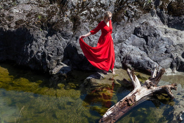 Блондинка в длинном красном платье на мистическом изображении парит в воздухе над зеленой водой озера на фоне каменного берега в горах Алтая. Магия и левитация. - Фото, изображение
