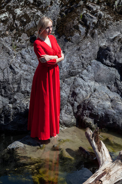 Uzun kırmızı elbiseli sarışın bir kadın, mistik bir görüntüyle gölün yeşil suyunun üzerinde Altai dağlarının arka planındaki taştan bir kıyıya doğru süzülüyor. Sihir ve havalanma. - Fotoğraf, Görsel