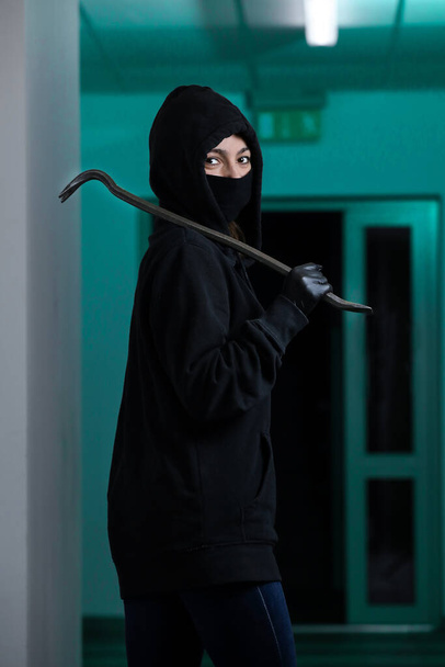 Thief broke into the apartment. House robbery by woman in a black jacket and black mask and crowbar. Burglar in a mask. Thief in a mask trying to break into other people's house - Fotoğraf, Görsel