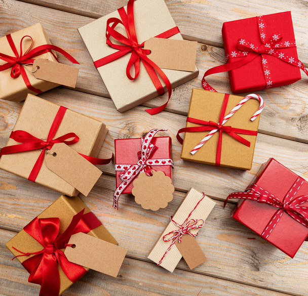 Christmas gift boxes with blank tags on wooden background. Flat lay of handmade present packages, kraft wrapping paper, red ribbon. Ideal for xmas celebration, festive event - Photo, Image