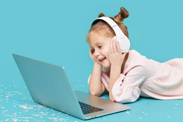 Smart child girl in wireless headphones using laptop on cyan background. Education, online study, tv, distance learning. Primary school student during quarantine. Educational games for children on a computer - Foto, Imagem