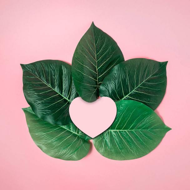 Pink greeting card with a heart-shaped paper in a frame of tropical green leaves. - Foto, immagini
