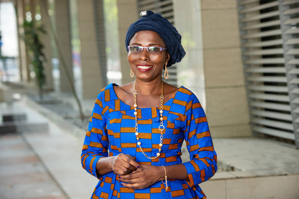 όμορφη Αφρικανή γυναίκα με παραδοσιακά ρούχα στέκεται σε εξωτερικούς χώρους κοιτάζοντας κάμερα χαμογελώντας. - Φωτογραφία, εικόνα
