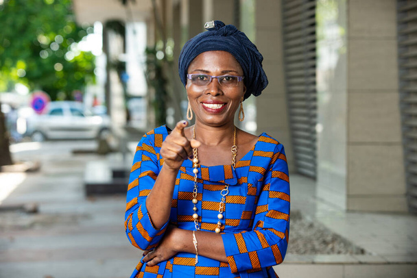 όμορφη Αφρικανή γυναίκα με παραδοσιακή στολή στέκεται σε εξωτερικούς χώρους δείχνοντας το δάχτυλο χαμογελώντας - Φωτογραφία, εικόνα