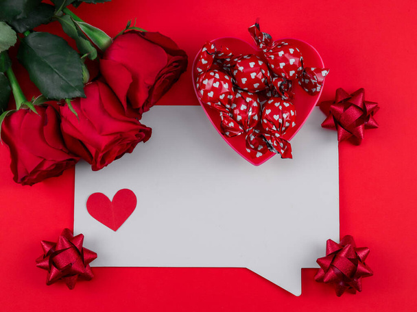 Weißes Blatt, drei Rosen, Schleife, Bonbons und ein Papierherz auf rotem Hintergrund mit Platz für Text in der Mitte, Nahaufnahme von oben. - Foto, Bild