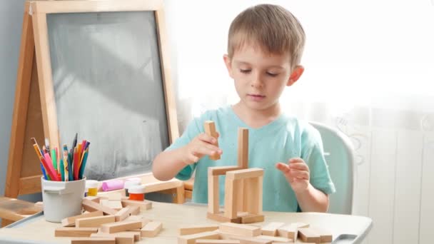 Portrét malého chlapce hrajícího si s dřevěnými tvárnicemi a stavební věží z cihel. Koncepce inteligentních dětí a domácí výchovy během uzamykání a vlastní izolace. - Záběry, video