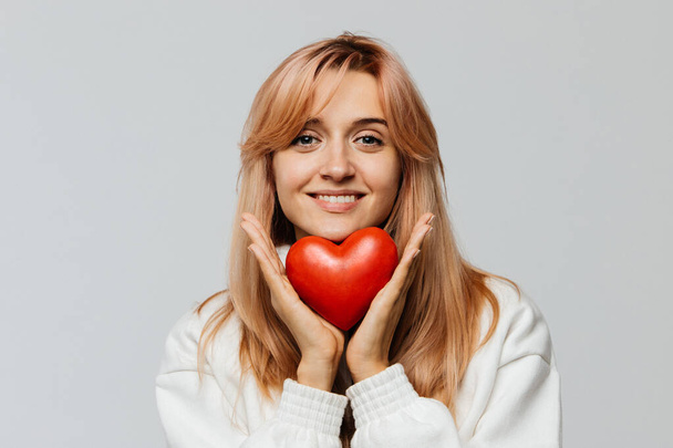 Πορτρέτο της χαρούμενης ελκυστική γυναίκα με φράουλα ξανθά μαλλιά κατέχουν κόκκινη καρδιά (σύμβολο του Αγίου Βαλεντίνου ημέρα), κοιτάζοντας κάμερα, closeup, απομονώνονται στο φως φόντο.Αγάπη, ευτυχία, σχέσεις έννοια - Φωτογραφία, εικόνα