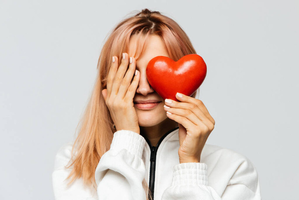 Happy cute young woman with strawberry blonde hair hold red heart (Valentine day symbol),covering her right eye with her palm, closeup, isolated on light background.Love, happiness concept - Photo, Image