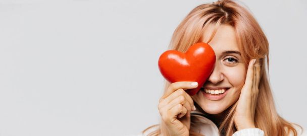 Portrait of joyful attractive woman with strawberry blonde hair hold red heart (Valentine day symbol),looking at camera, closeup, isolated on light background.Love, happiness, relationships concept - Foto, imagen