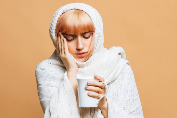 Sickness cute young woman with strawberry blonde hair, warm plaid with headache, holding cup of hot tea, touching her temple, feeling the first symptoms of illness. Flu, feeling unwell, cold concept - Photo, Image