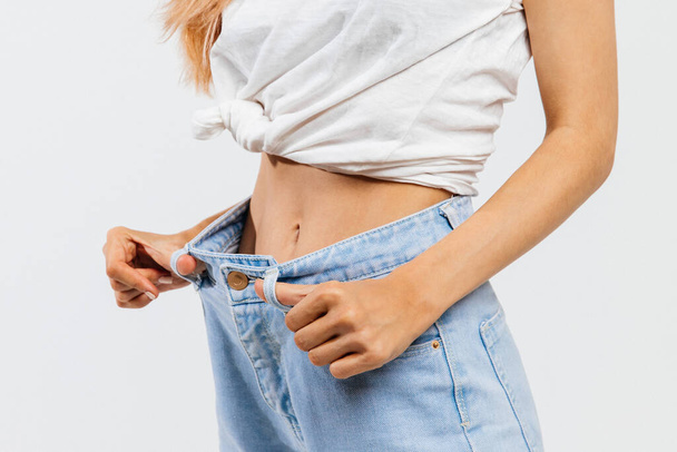 Diet, active sports training, slim waist concept. Closeup of woman in loose blue jeans showing/demonstrating successful weight loss, isolated on white background. - Fotografie, Obrázek