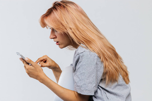 Έκπληκτος σύγχυση νεαρή γυναίκα με μακριά μαλλιά χρησιμοποιώντας και κοιτάζοντας smartphone / κινητό τηλέφωνο, απομονώνονται σε γκρι φόντο, πλευρική άποψη. Συγκλονιστικό μήνυμα, άσχημα νέα.. - Φωτογραφία, εικόνα