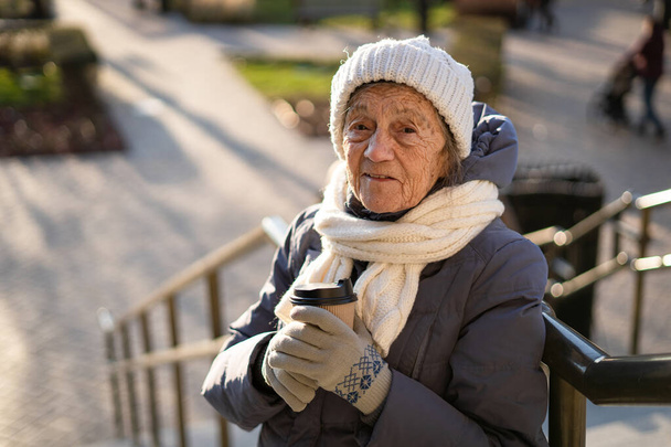 Portret szczęśliwej starszej białej kobiety z filiżanką kawy w zimowych ciuchach, białą dzianiną i szalikiem w słonecznym mieście pogodowym. Starsza pani z kawą na świeżym powietrzu. Podróże, zabawa, emerytura - Zdjęcie, obraz