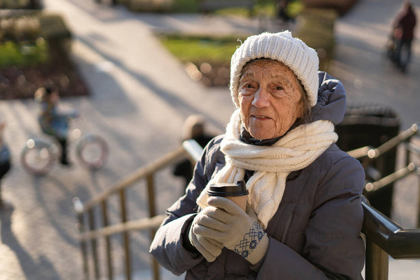 Χαριτωμένη ηλικιωμένη γυναίκα στην πόλη πίνει καφέ με τον εαυτό της σε χειμερινό παλτό και ζεστό καπέλο, κασκόλ και πλεκτά γάντια σε ηλιόλουστο χειμώνα καιρό. Όμορφη γριά με χάρτινο φλιτζάνι πρωινό καφέ για έξω.. - Φωτογραφία, εικόνα