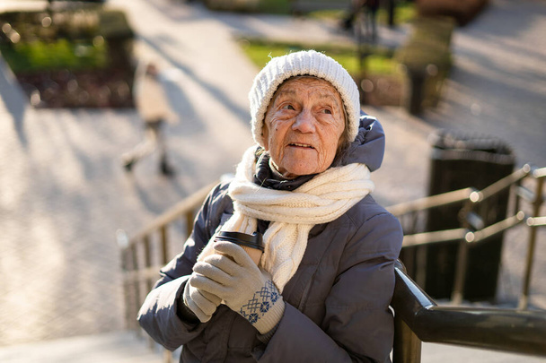 Słodka starsza kobieta w mieście pije kawę ze sobą w zimowym płaszczu i ciepłym kapeluszu, szaliku i rękawiczkach na drutach w słoneczną zimową pogodę. Przystojna staruszka z papierową filiżanką porannej kawy.. - Zdjęcie, obraz