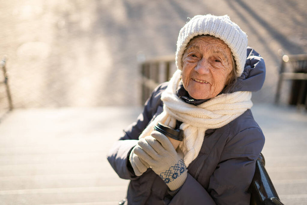 Χαριτωμένη ηλικιωμένη γυναίκα στην πόλη πίνει καφέ με τον εαυτό της σε χειμερινό παλτό και ζεστό καπέλο, κασκόλ και πλεκτά γάντια σε ηλιόλουστο χειμώνα καιρό. Όμορφη γριά με χάρτινο φλιτζάνι πρωινό καφέ για έξω.. - Φωτογραφία, εικόνα