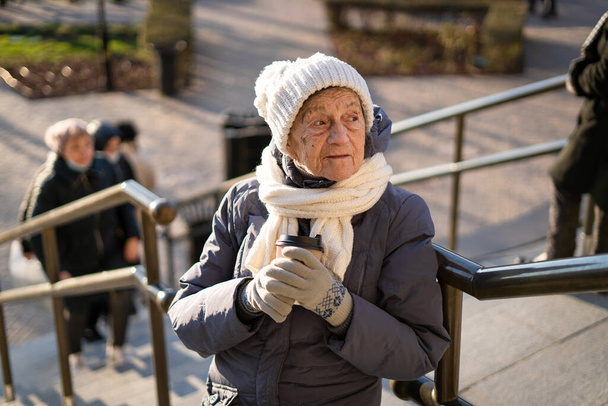 Vidám idős hölgy kezében kávé elvitelre, öltözött téli ruhák a városban. Gyönyörű idős hölgy egy csésze kávéval elvitelre. Aktív pihenés idős korban, kávészünet séta közben. Forró ital, nyugdíjas 90 év. - Fotó, kép