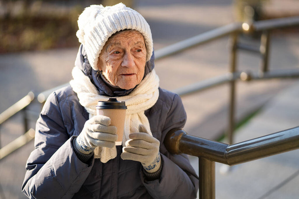 Χαρούμενη ηλικιωμένη κυρία που κρατάει καφέ για να φύγει, ντυμένη χειμωνιάτικα ρούχα στην πόλη. Όμορφη ηλικιωμένη κυρία με καφέ για έξω. Ενεργός ανάπαυση σε μεγάλη ηλικία, διάλειμμα για καφέ κατά τη διάρκεια του περπατήματος. Ζεστό ποτό, συνταξιούχος 90 χρόνια. - Φωτογραφία, εικόνα