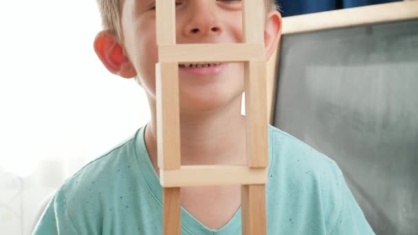 Szczęśliwy śmiech i uśmiech chłopca patrząc przez wysoką drewnianą wieżę z bloków i cegieł. Koncepcja edukacji dzieci w domu podczas zamknięcia i pozostawania w domu - Materiał filmowy, wideo