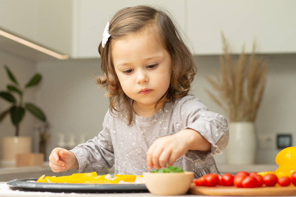 Nettes kleines Mädchen 2-4 in grauem Kleid, das in der Küche Pizza kocht. Kind arrangiert Zutaten auf Pizzaboden - Foto, Bild