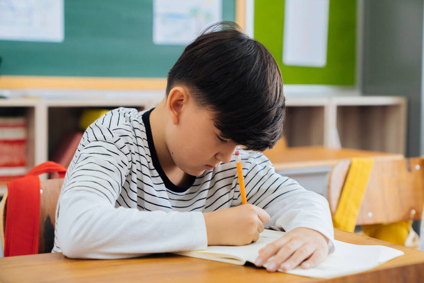 Caucasian school boy sitting in school writing in book with pencil, studying, education, learning. Male student sitting at desk in classroom writing in notebook in exam - Фото, изображение