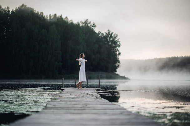 Happy woman in a white dress enjoys morning on a lake. Fog floats over the water. - Фото, изображение