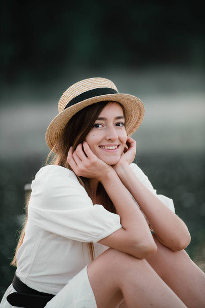 Portret van een gelukkige blanke vrouw in een witte jurk en hoed buiten zittend en glimlachend. - Foto, afbeelding