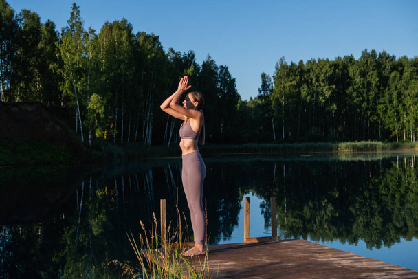 Giovane donna sul lago che pratica yoga si muove su una piattaforma di legno. Bella giovane donna che si esercita in natura, stile di vita sano giovani vibrazioni positive. - Foto, immagini