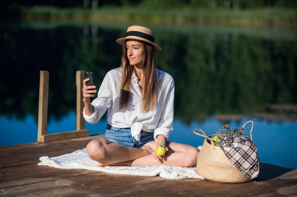 Schöne junge Frau mit Hut picknickt in der Nähe des Sees im Sommerwald. Sie isst einen Apfel und schaut aufs Smartphone. - Foto, Bild
